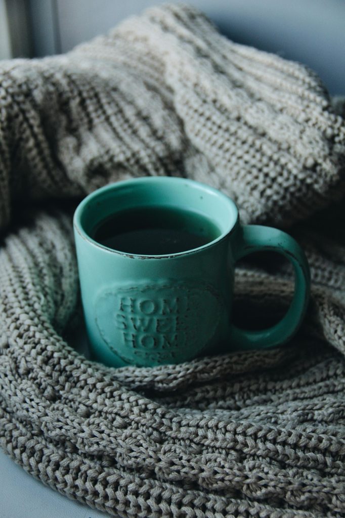 Türkise Teetasse mit der Prägung „Home Sweet Home“ liegt in eine Strickdecke gehüllt – Sinnbild für Wärme, Geborgenheit und Rückzug im Winter